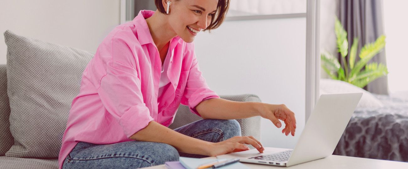 Na imagem temos uma mulher branca sentada num sofá cinza enquanto mexe em seu notebook branco. Ela tem um cabelo curto e castanho e usa fones de ouvido sem fio. Sorrindo, simboliza o benefício de uma internet rápida.