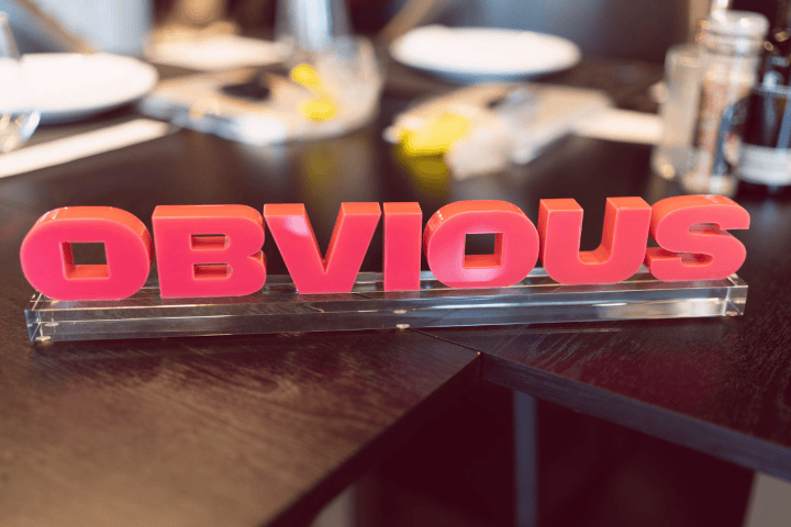 Placa da logo da empresa OBVIOUS em 3D, sobre uma mesa de escritório