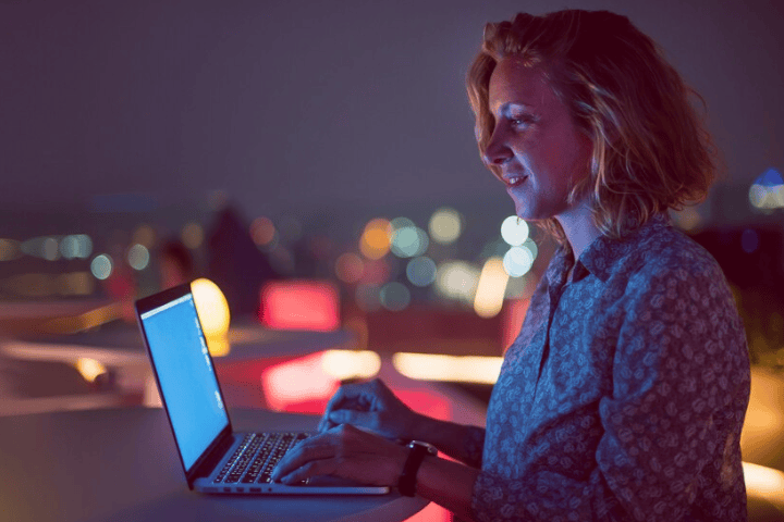 Mulher mexendo em seu notebook no topo de um prédio, com vista para as luzes da cidade